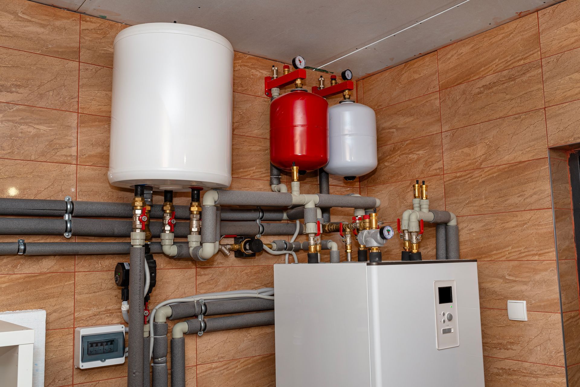 White water heater and auxiliary tanks, pipes with insulation, and a control unit against a tiled wall.