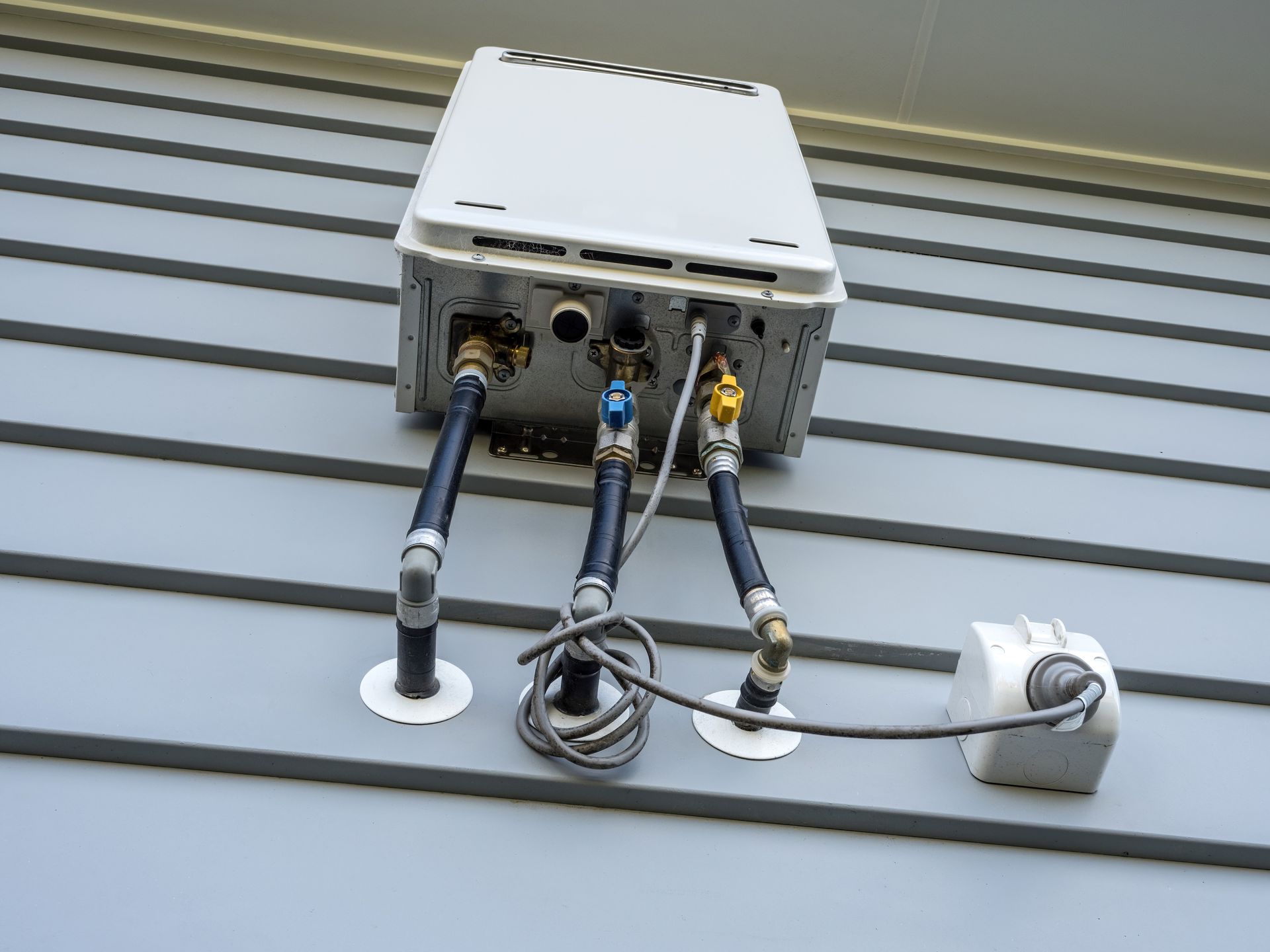 White tankless water heater mounted on gray siding with three pipes and a connected electrical box.