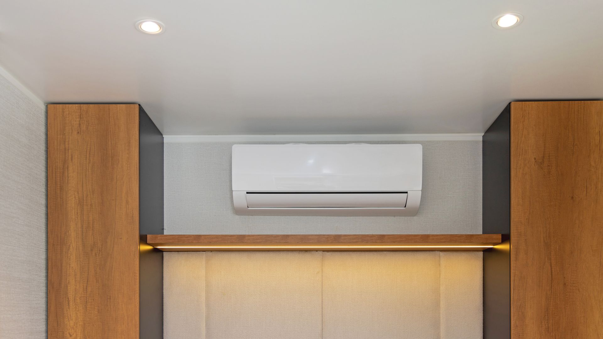 White air conditioner mounted on wall, flanked by wood-paneled cabinets, with recessed lighting.
