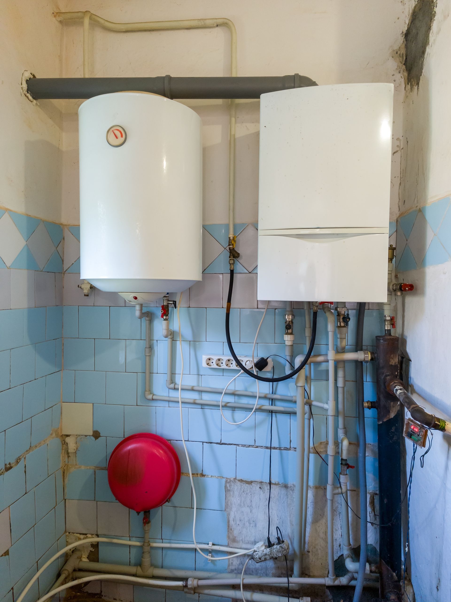 White water heater and boiler on a blue-tiled wall with pipes and a red expansion tank.
