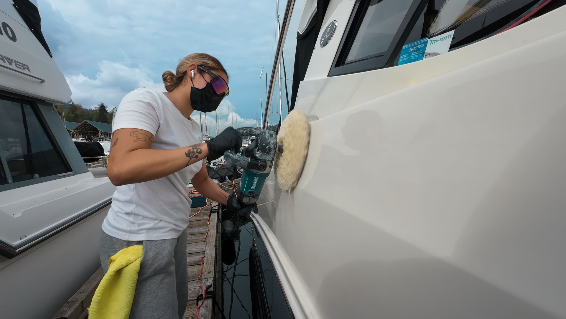 Person using a power buffer to polish a boat's white hull at a marina; wearing a mask, safety glasses, and gloves.