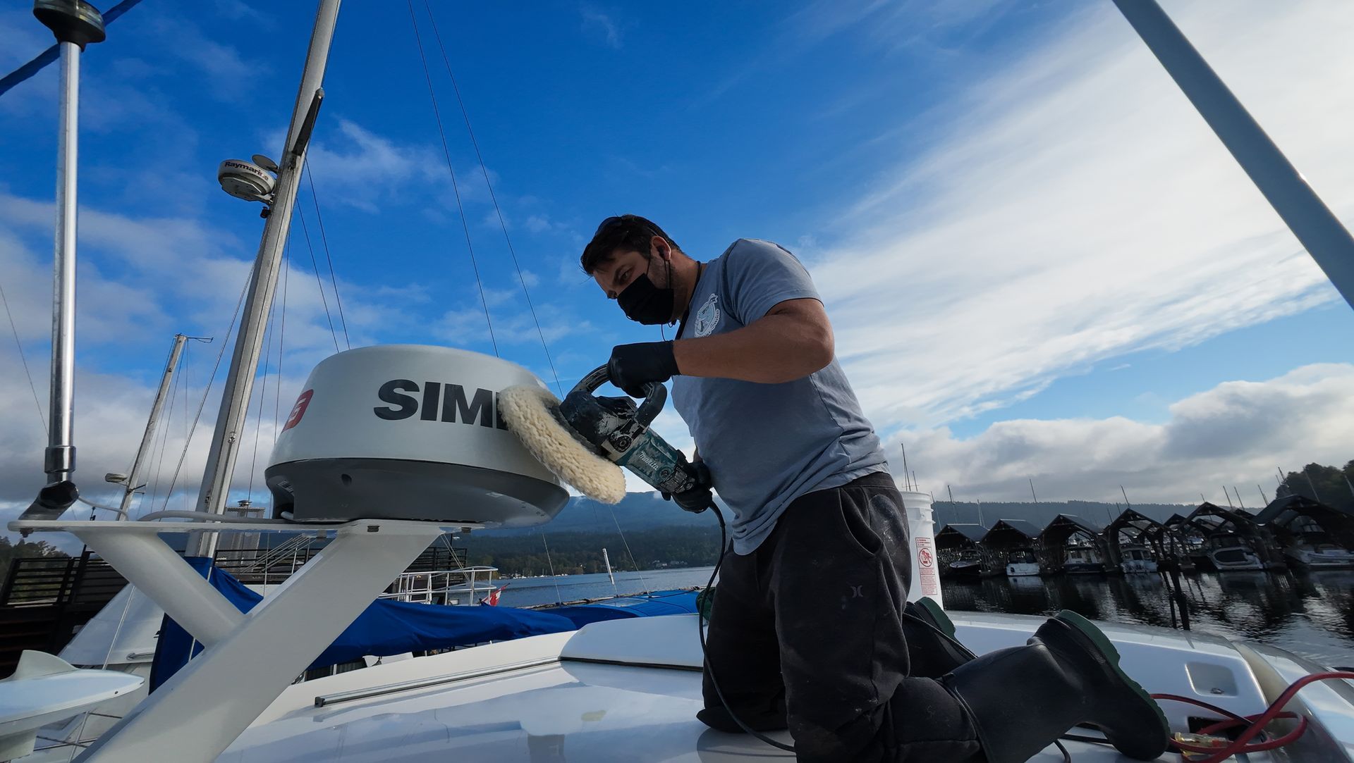 Man kneeling on boat deck, sanding radar dome, wearing mask and gloves. Bright blue sky.