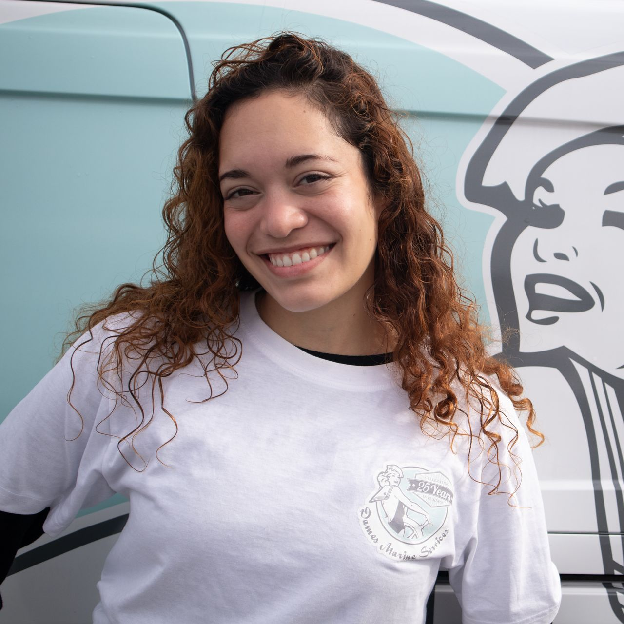 Woman with curly brown hair smiles, wearing a white t-shirt. Standing beside a light blue van with a graphic.