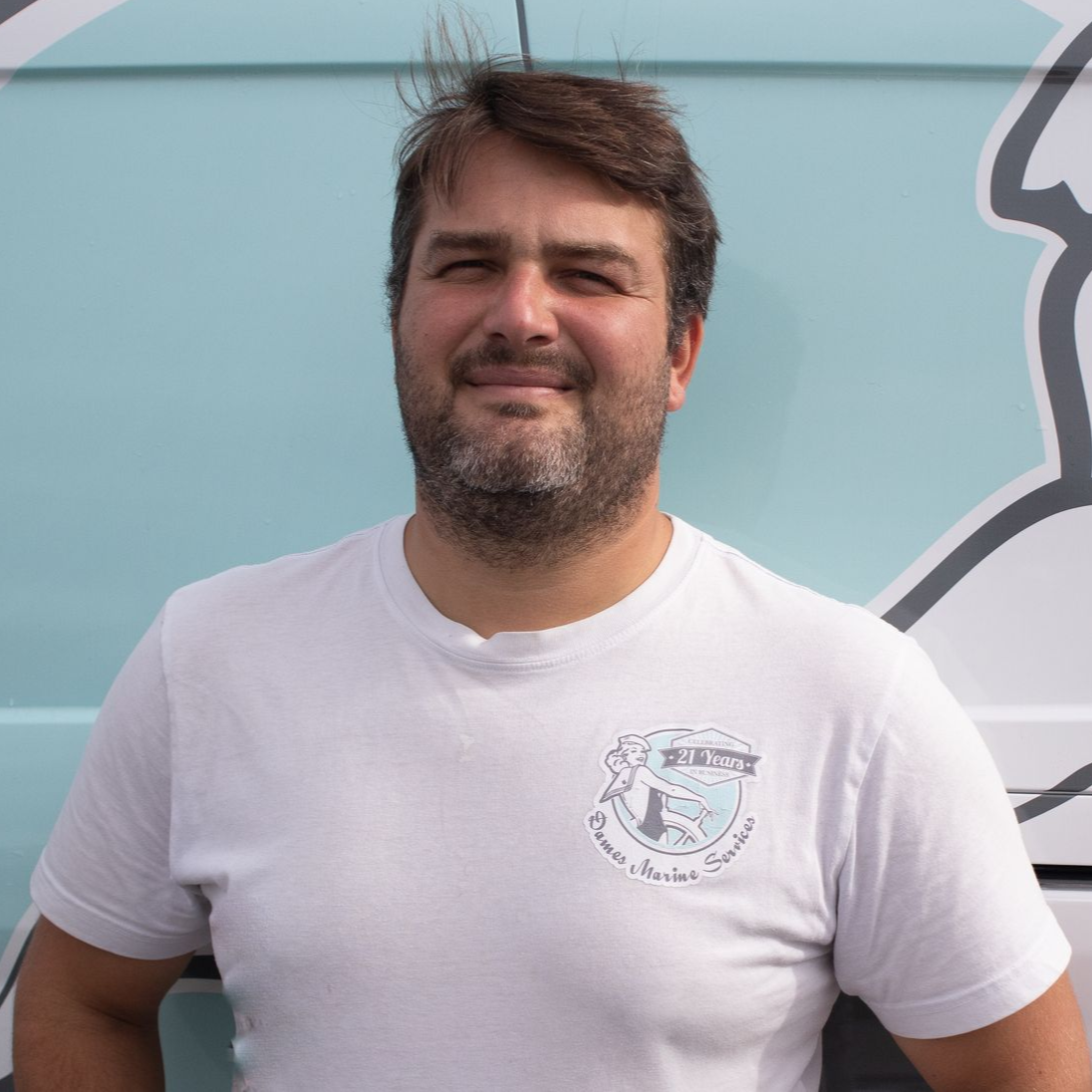 Man in white shirt with logo smiles, stands in front of light blue van.