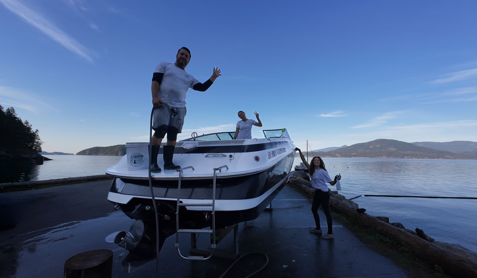 Three people on a boat by the water, waving. Bright blue sky.