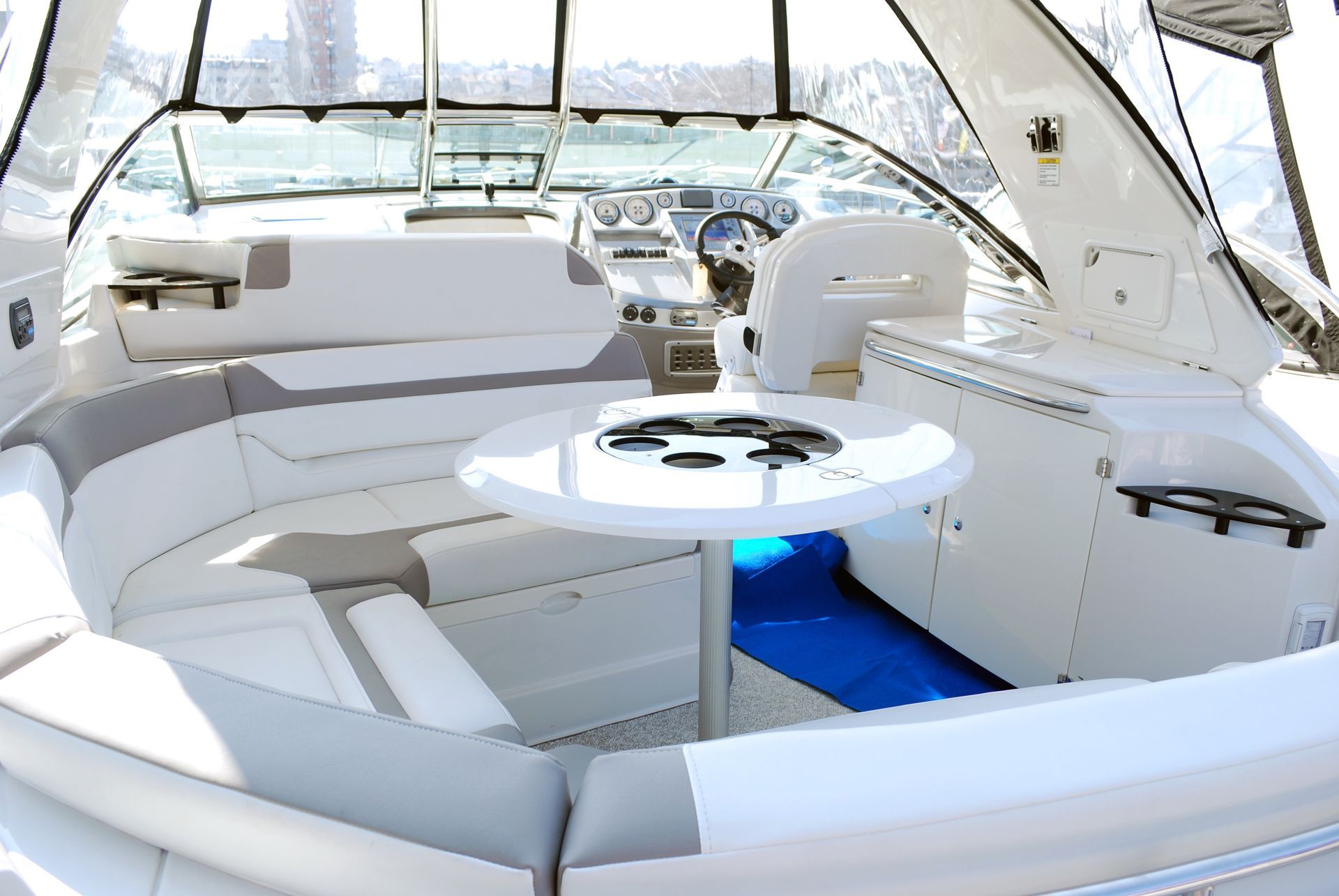 Interior of a white boat with circular table and seating. Blue accents on cushions. Clear windshield.