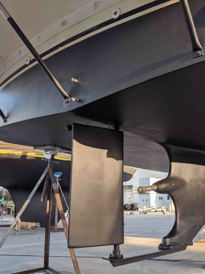 Black sailboat hull with keel and rudder visible, supported on jacks at a shipyard.