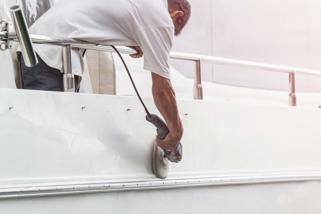 Person using a power tool to sand the side of a white boat near a railing.