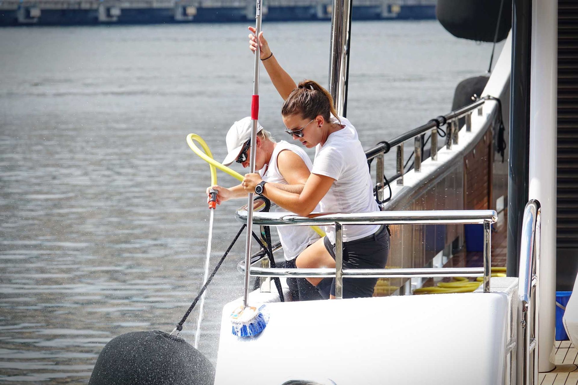 Two people washing a yacht with a hose. One person is holding a pole. The water is spraying.