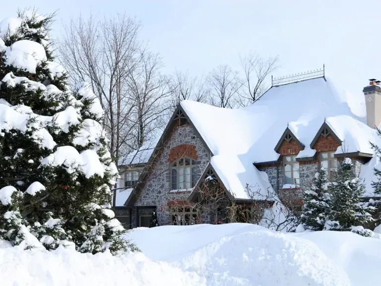 Snow-covered stone house with a steep roof, next to a snow-laden evergreen tree, under a bright, winter sky.