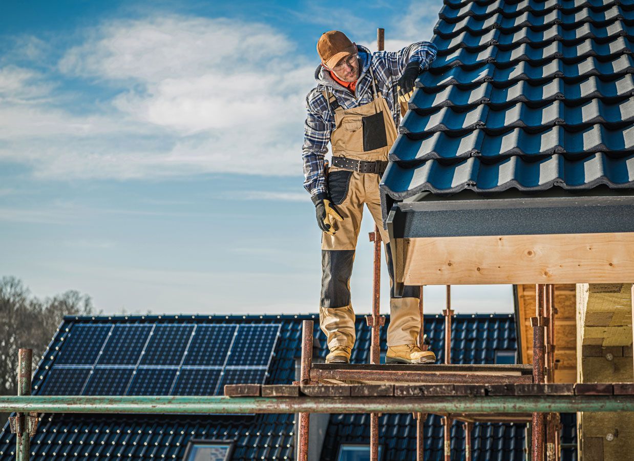 Roofer on scaffolding installing dark roof tiles on a sunny day. Solar panels visible on another roof.