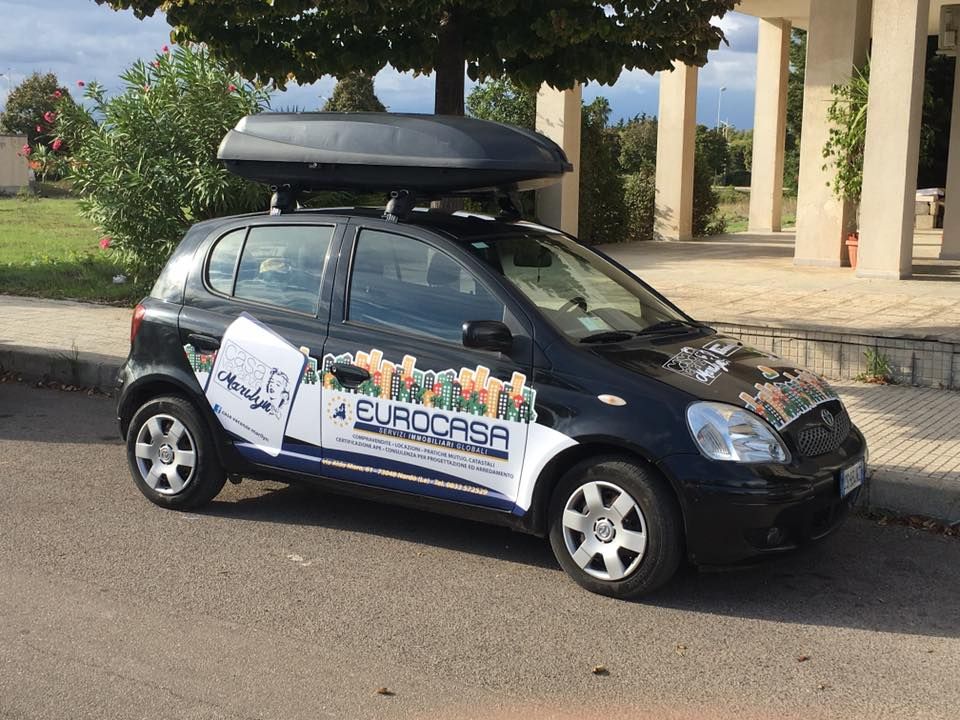 Un'auto nera con un box sul tetto è parcheggiata sul ciglio della strada.