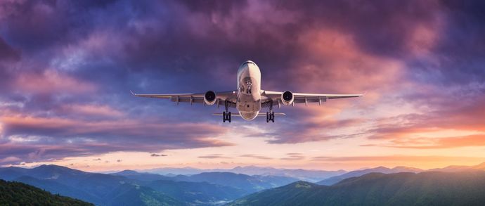 A passenger airplane flying head-on over a mountain range during a vibrant, purple and orange sunset.