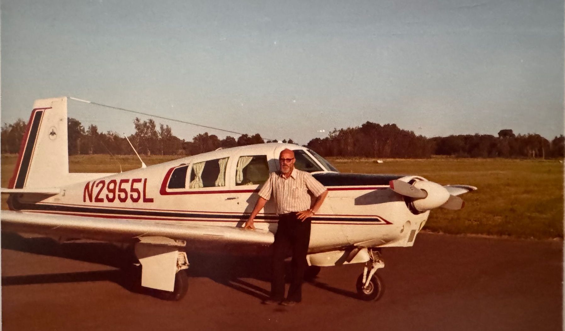 A man standing beside a small white airplane with red and blue stripes and registration number N2955L on a tarmac.