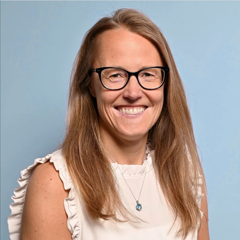 Smiling person with long light brown hair and black-rimmed glasses, wearing a white ruffled top against a blue background.