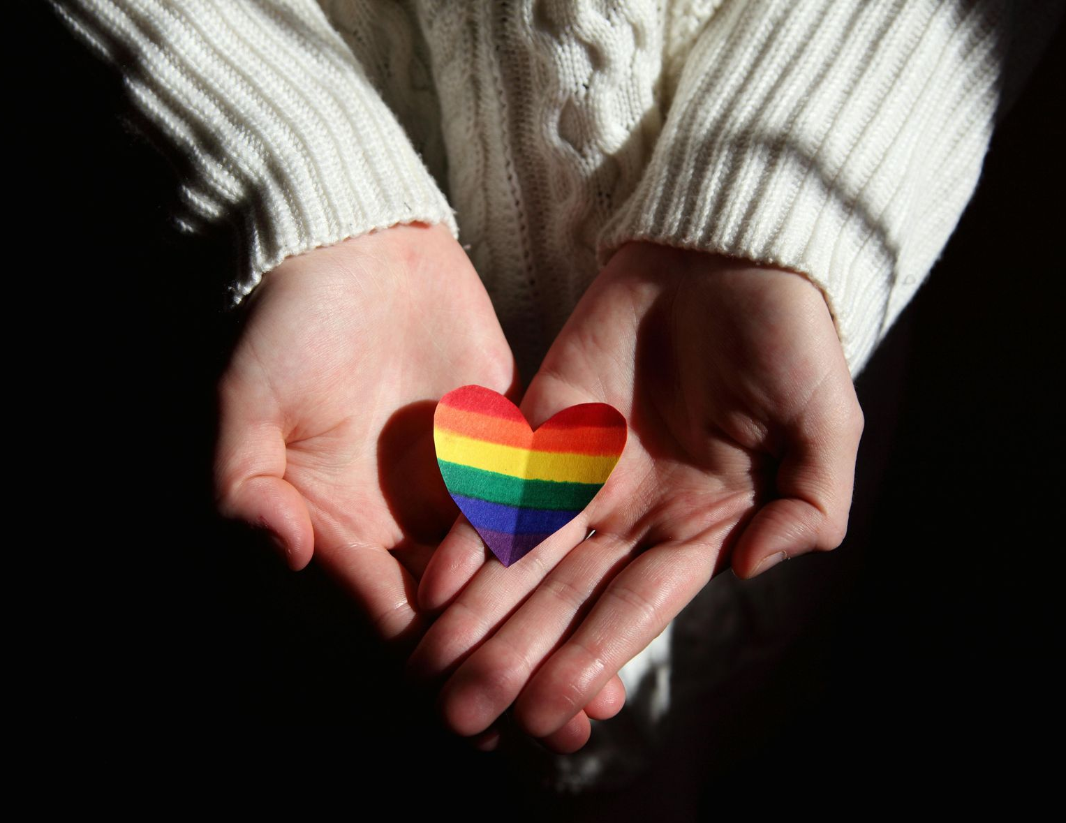 A person wearing a white sweater holds a small paper heart with rainbow stripes in their cupped hands.