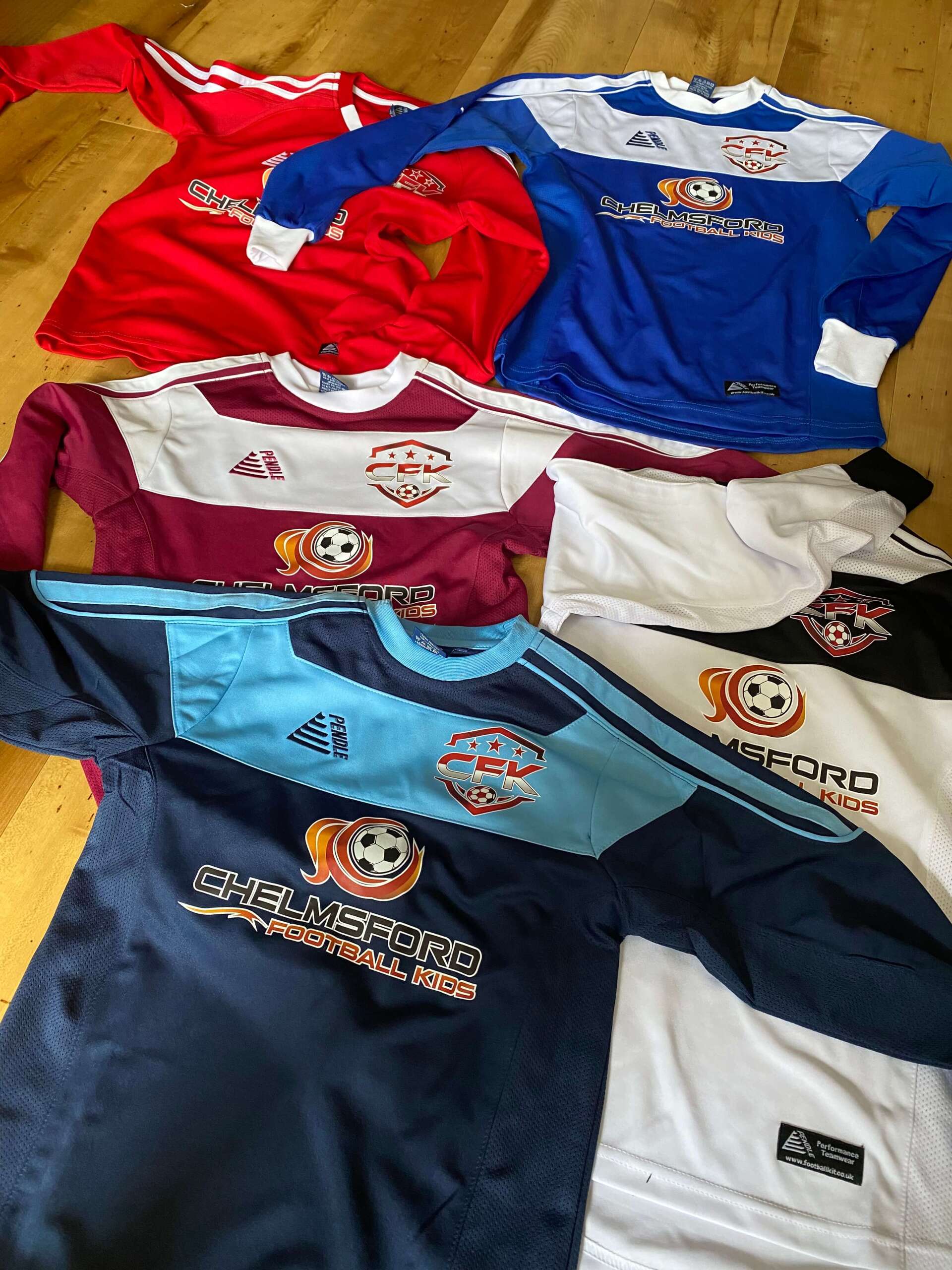 Six colorful soccer jerseys in red, blue, maroon, white, and navy blue, with logos and sponsor details.