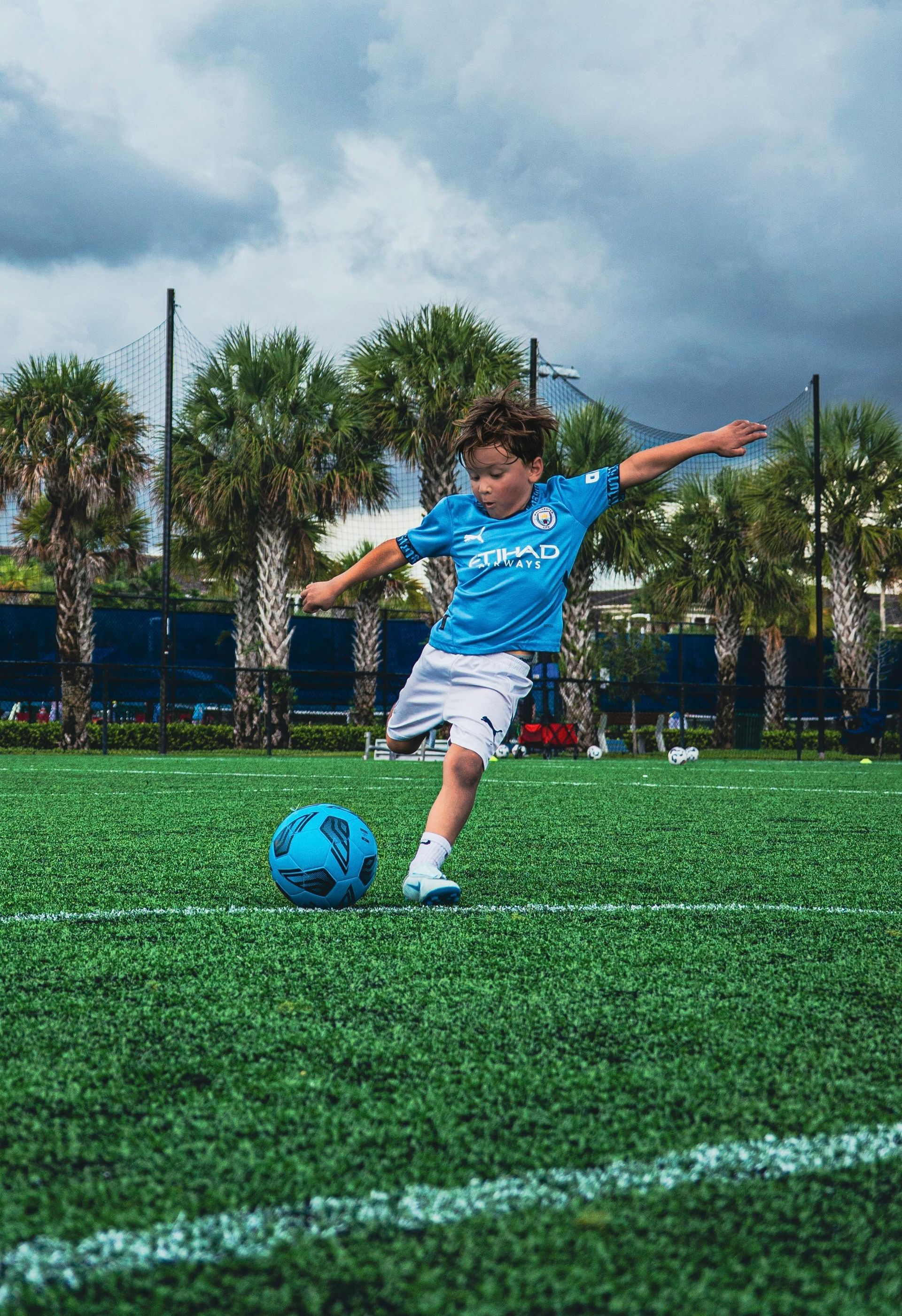 Young boy in blue uniform kicking a soccer ball on a green field under a cloudy sky.