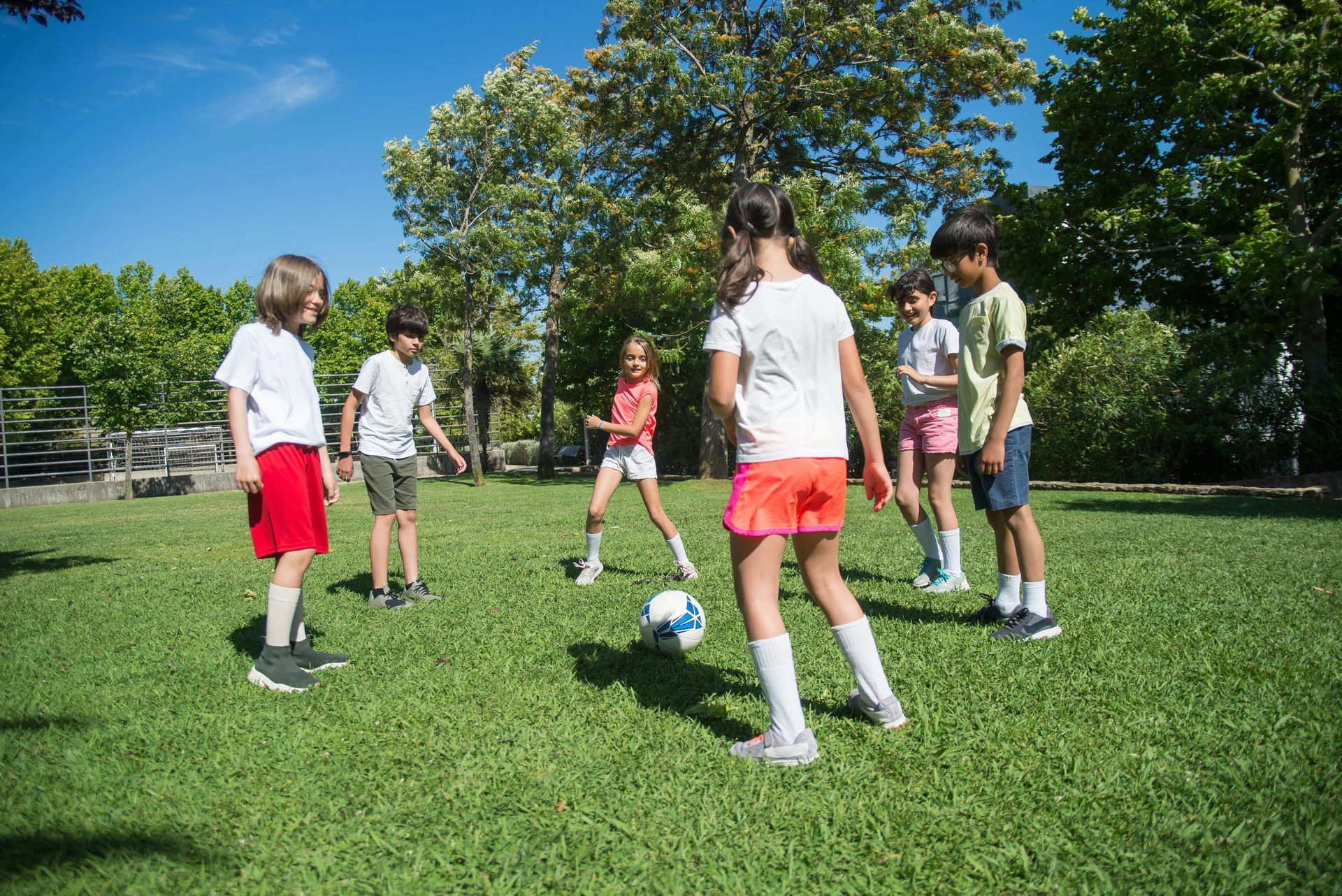 Children playing soccer on a grassy field in bright sunlight; some wear red and orange shorts.