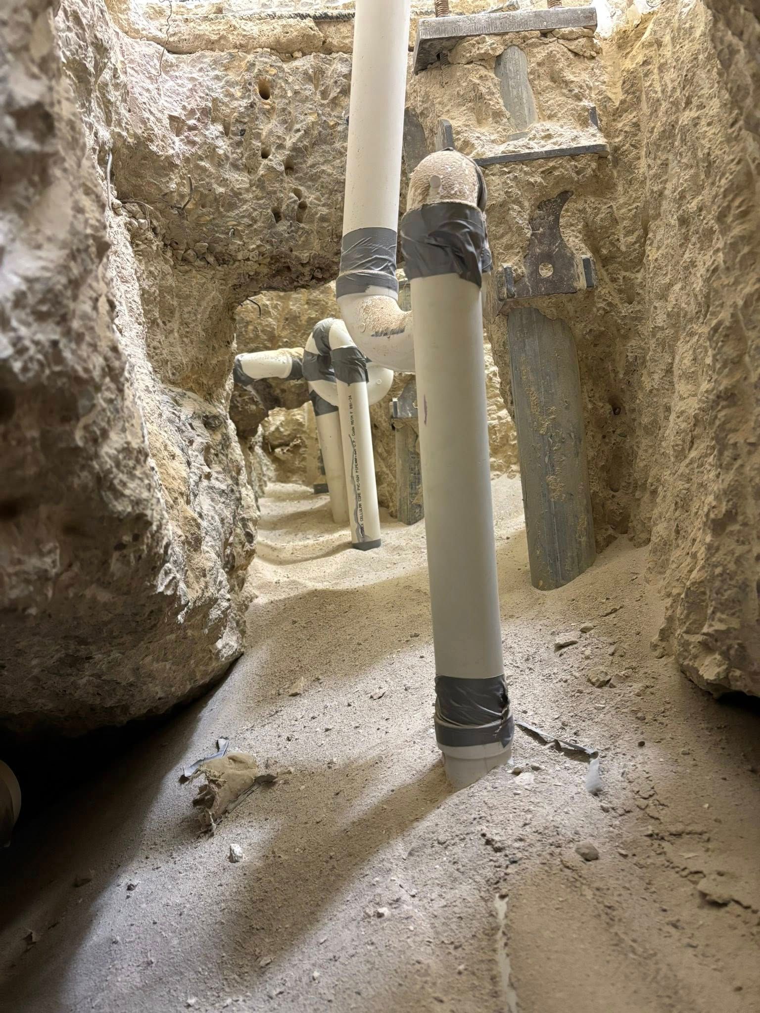 PVC plumbing pipes installed upright in a trench filled with light-colored sand and rocky, excavated soil.
