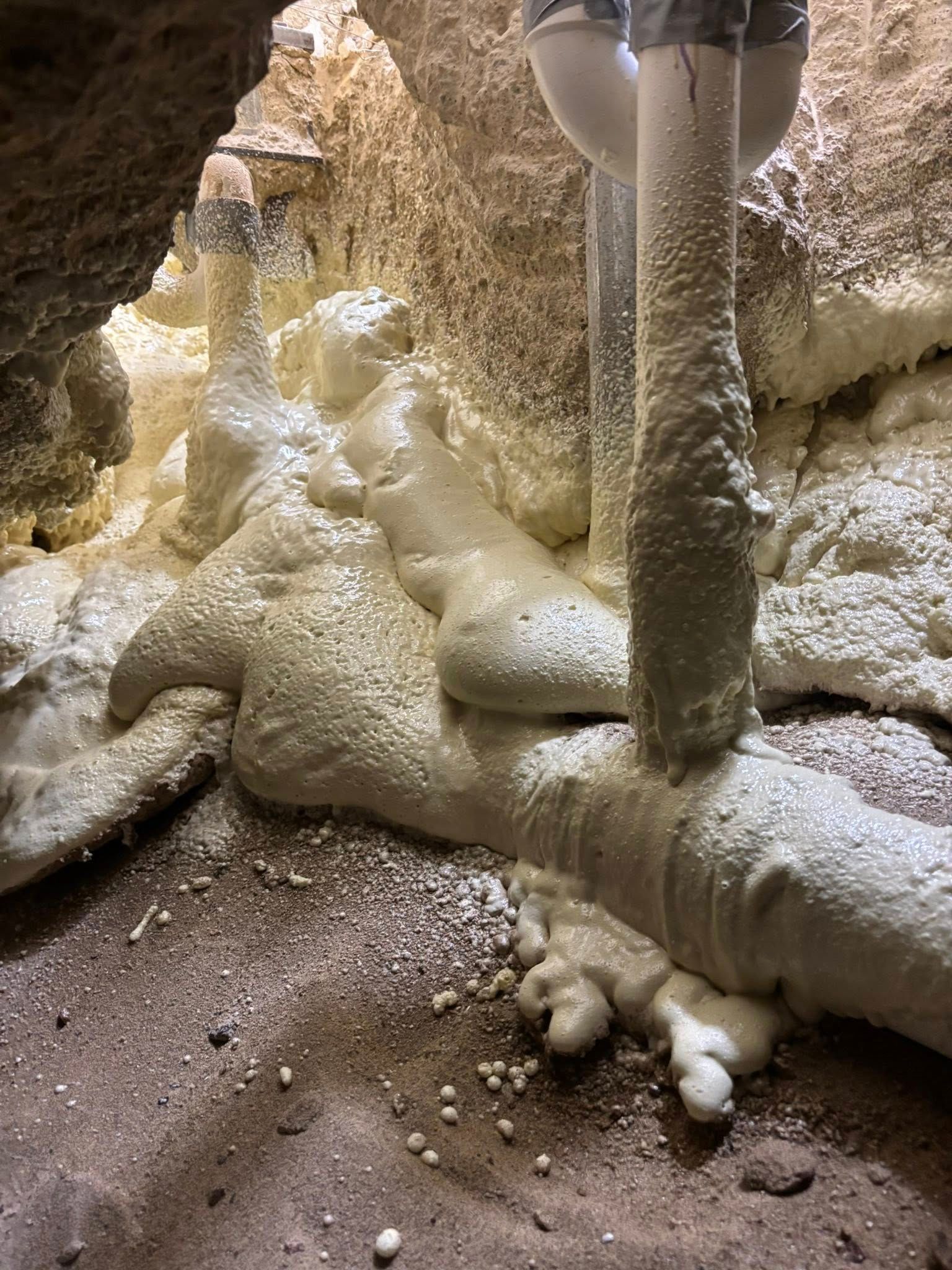 Expanding yellow spray foam fills a dark crawl space, surrounding pipes and covering the dirt ground.