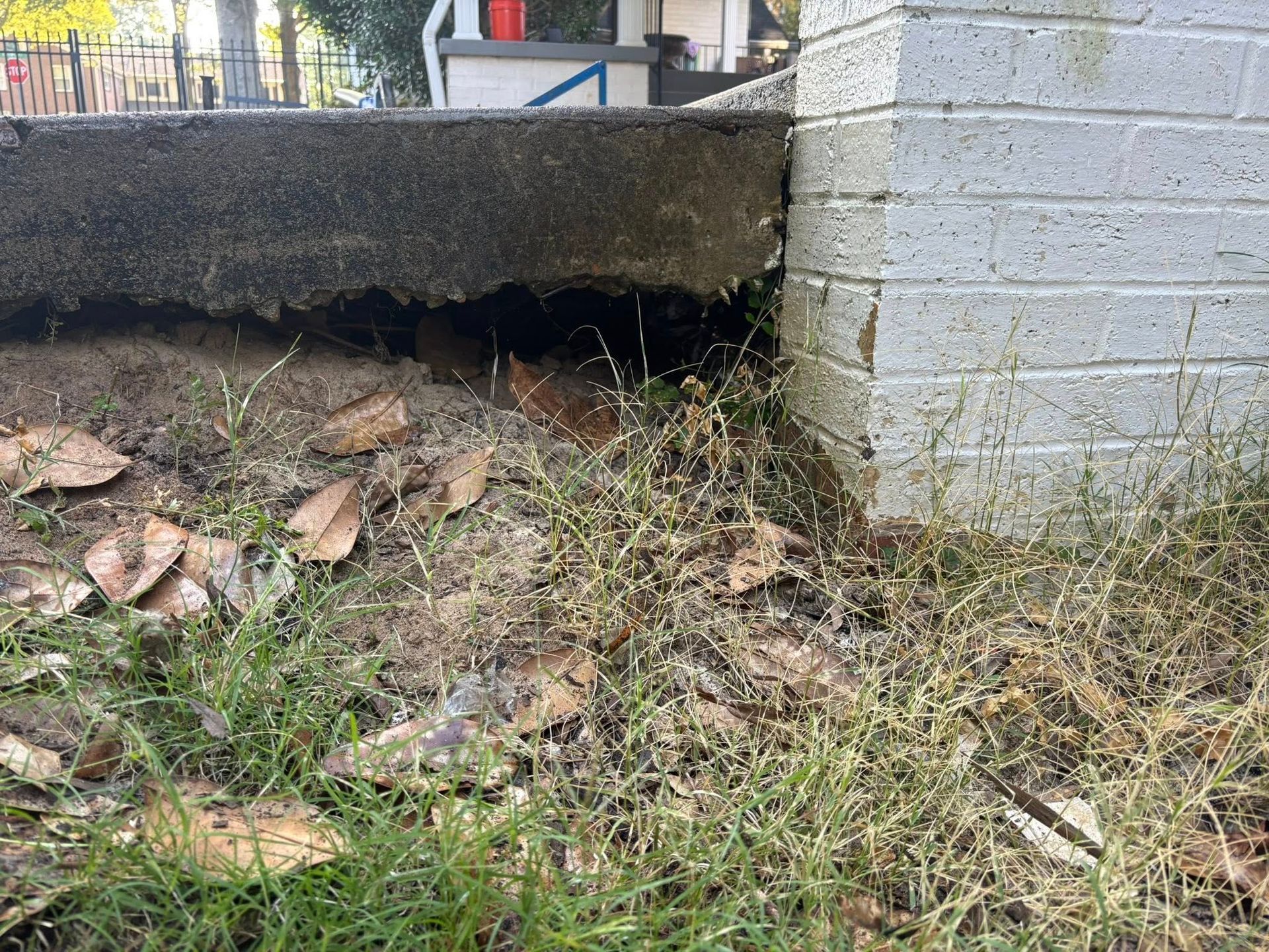 A concrete slab showing significant soil erosion and a large gap beneath the edge next to a white brick wall.
