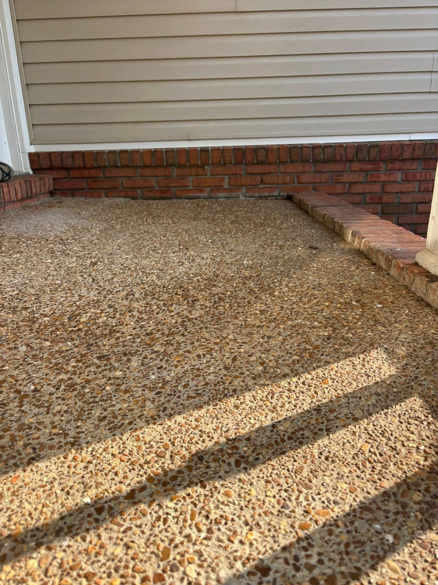 An outdoor porch with a textured pebble-concrete floor, bordered by a red brick foundation and light-colored siding.