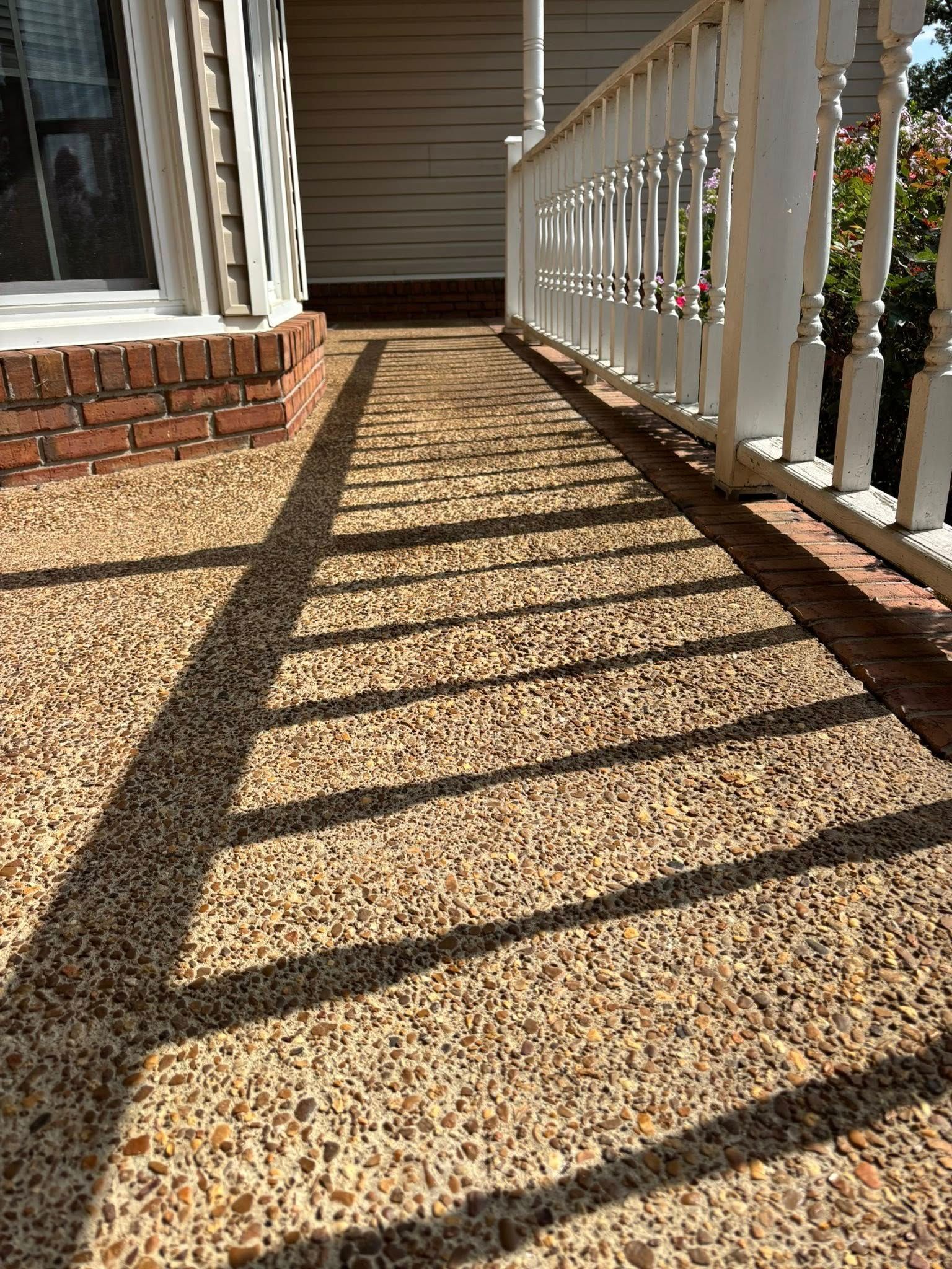 A sunlit porch with a pebble-textured floor, bordered by brick and a white railing, casting long, distinct shadows.
