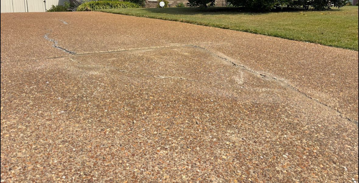 A brown, textured concrete driveway with a light, jagged surface crack near a patch of grass.