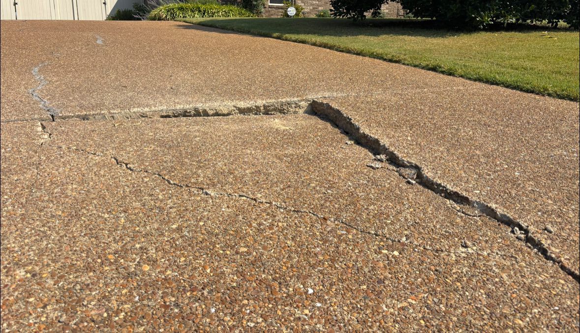 A damaged outdoor concrete driveway showing a deep, rectangular crack and surface separation.