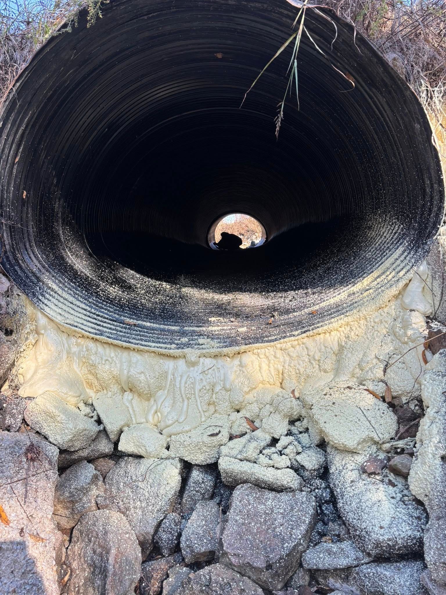 View looking into a large, dark, corrugated metal culvert pipe set into a base of light-colored, rocky cement.