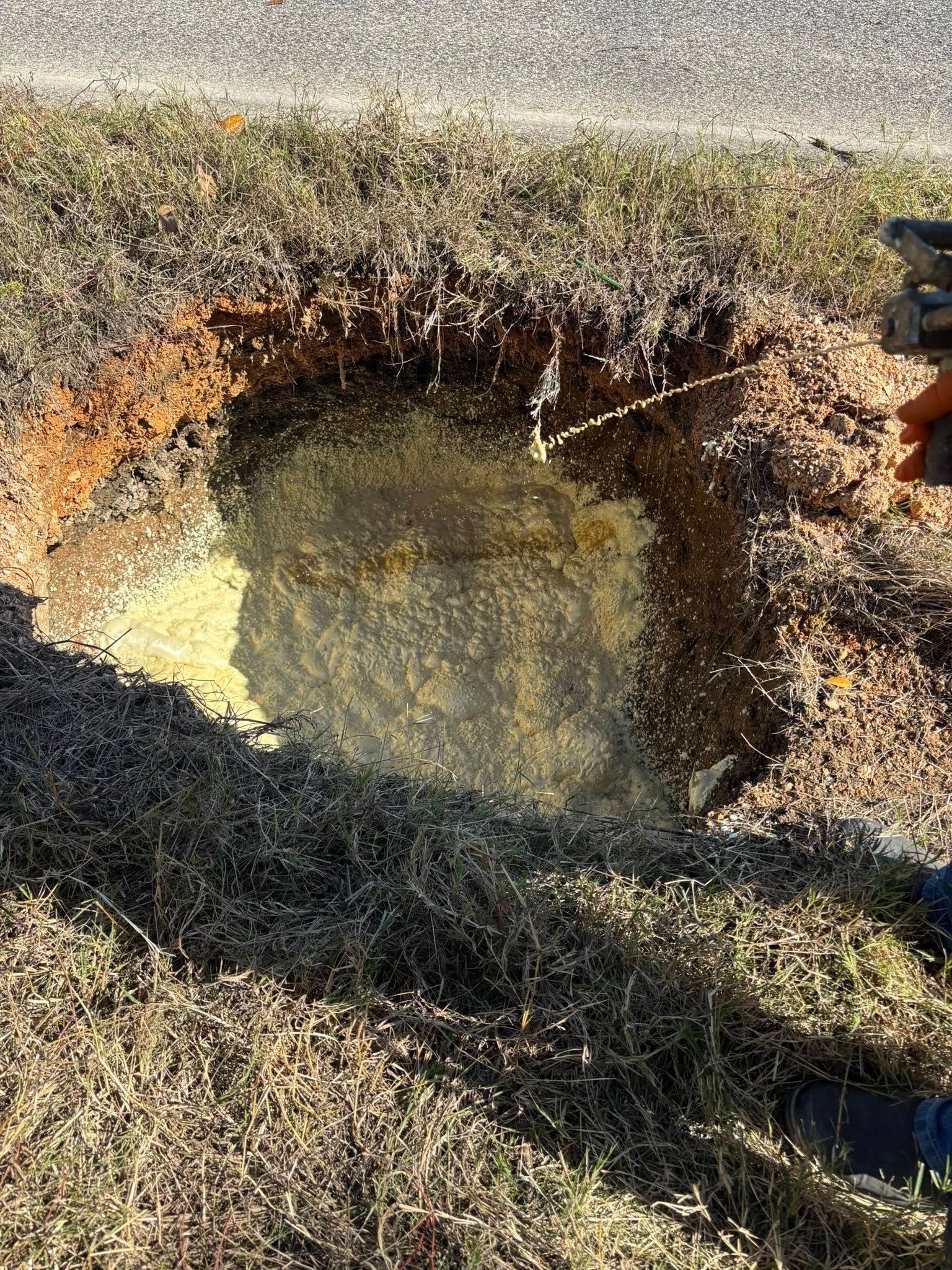 A circular hole in the grassy roadside revealing yellowish soil and a partially visible pipe or utility line beneath.