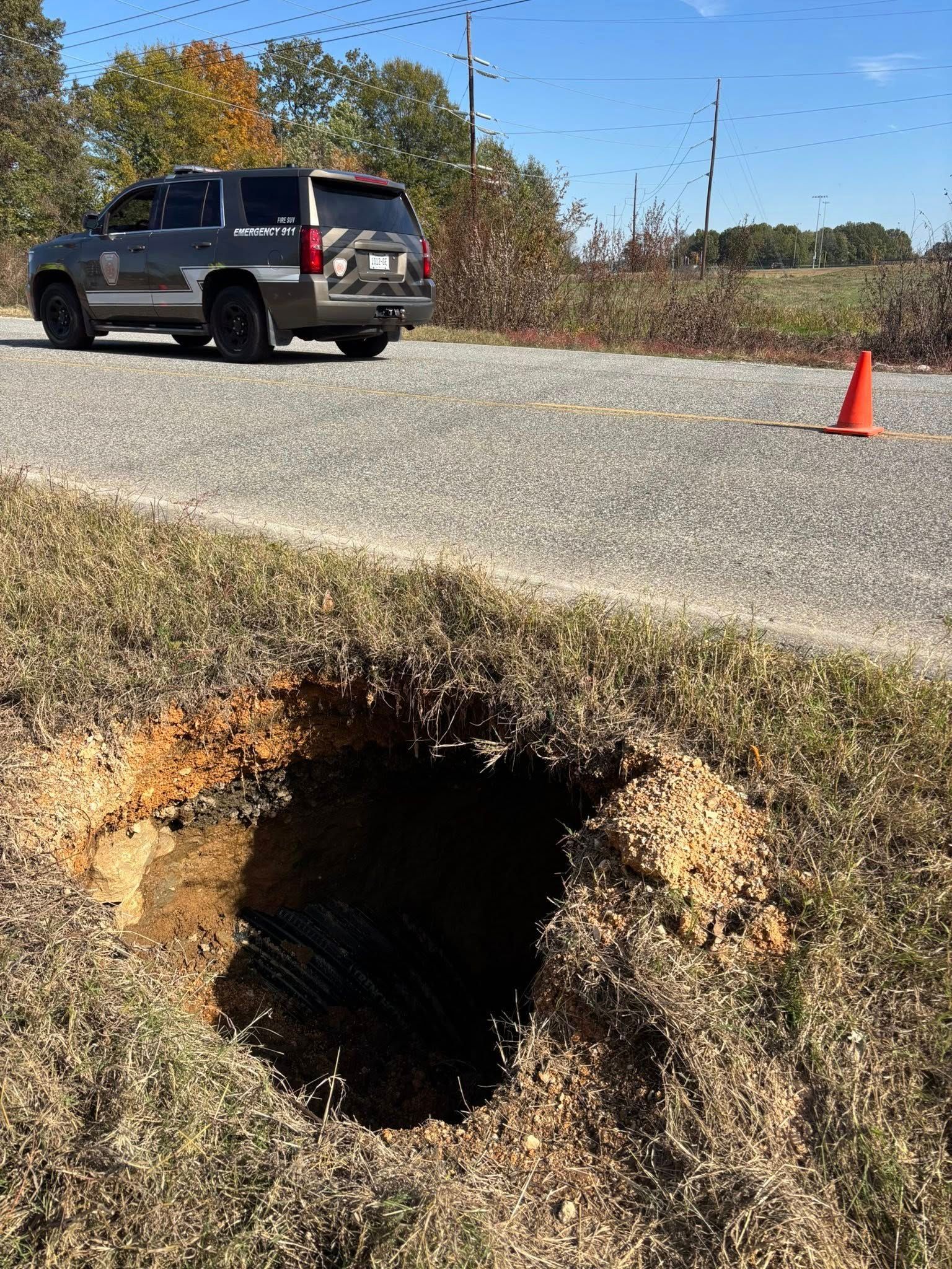 A large sinkhole in a grassy roadside area, with a parked law enforcement vehicle and a traffic cone in the background.
