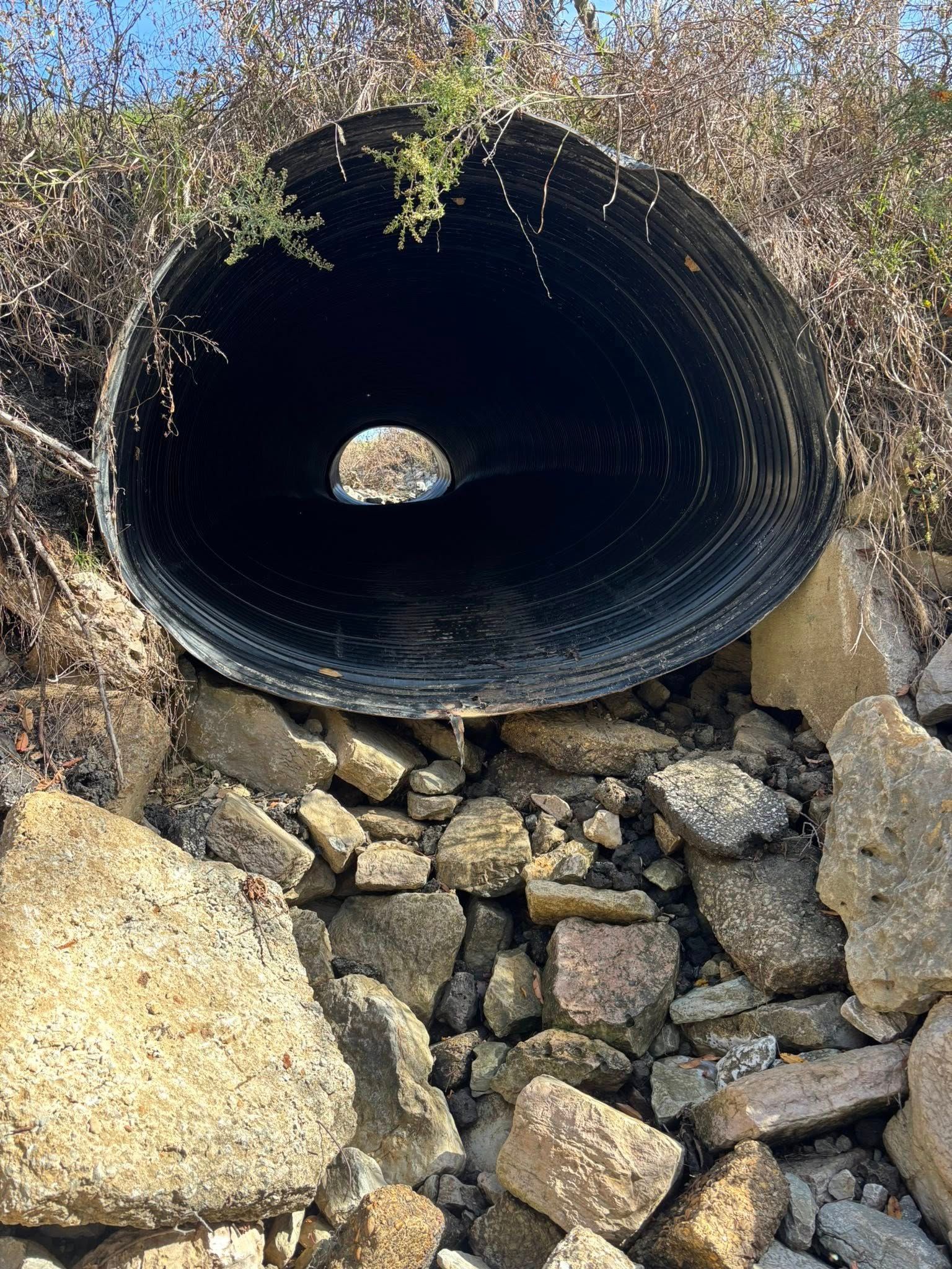 A large, circular corrugated metal drainage pipe embedded in a rocky, overgrown embankment, viewed from the opening.