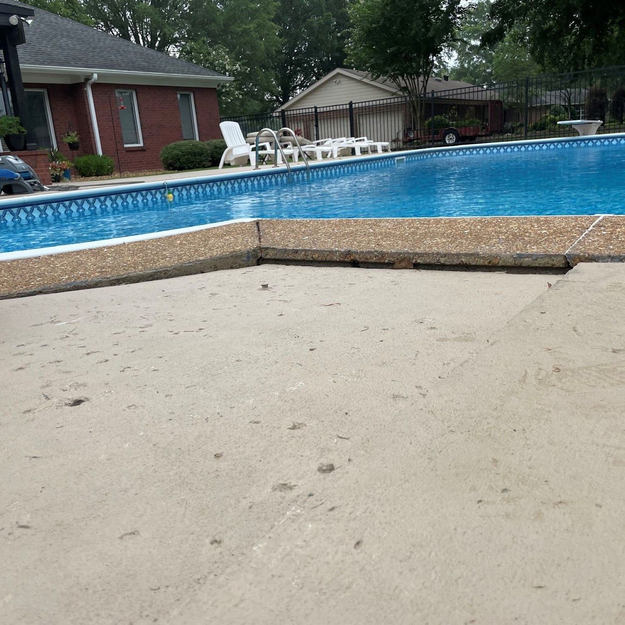 A swimming pool with blue water and a textured concrete deck, set beside a red brick house and a white garage.