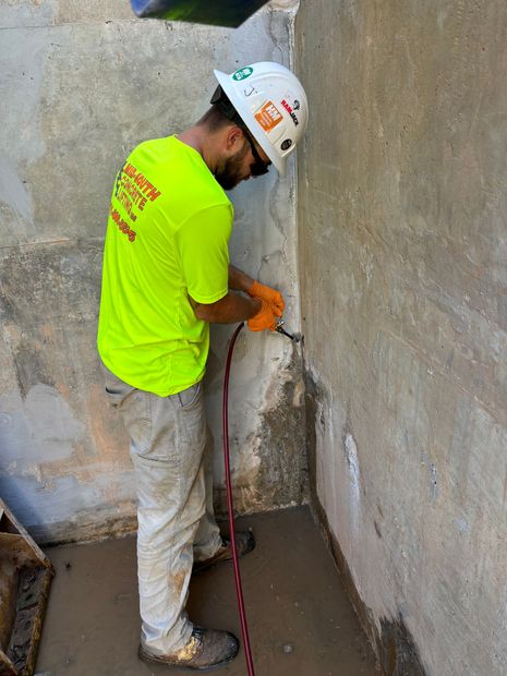 A person uses an electric drill to create a hole in a concrete surface, with a small metal anchor and wrench nearby.