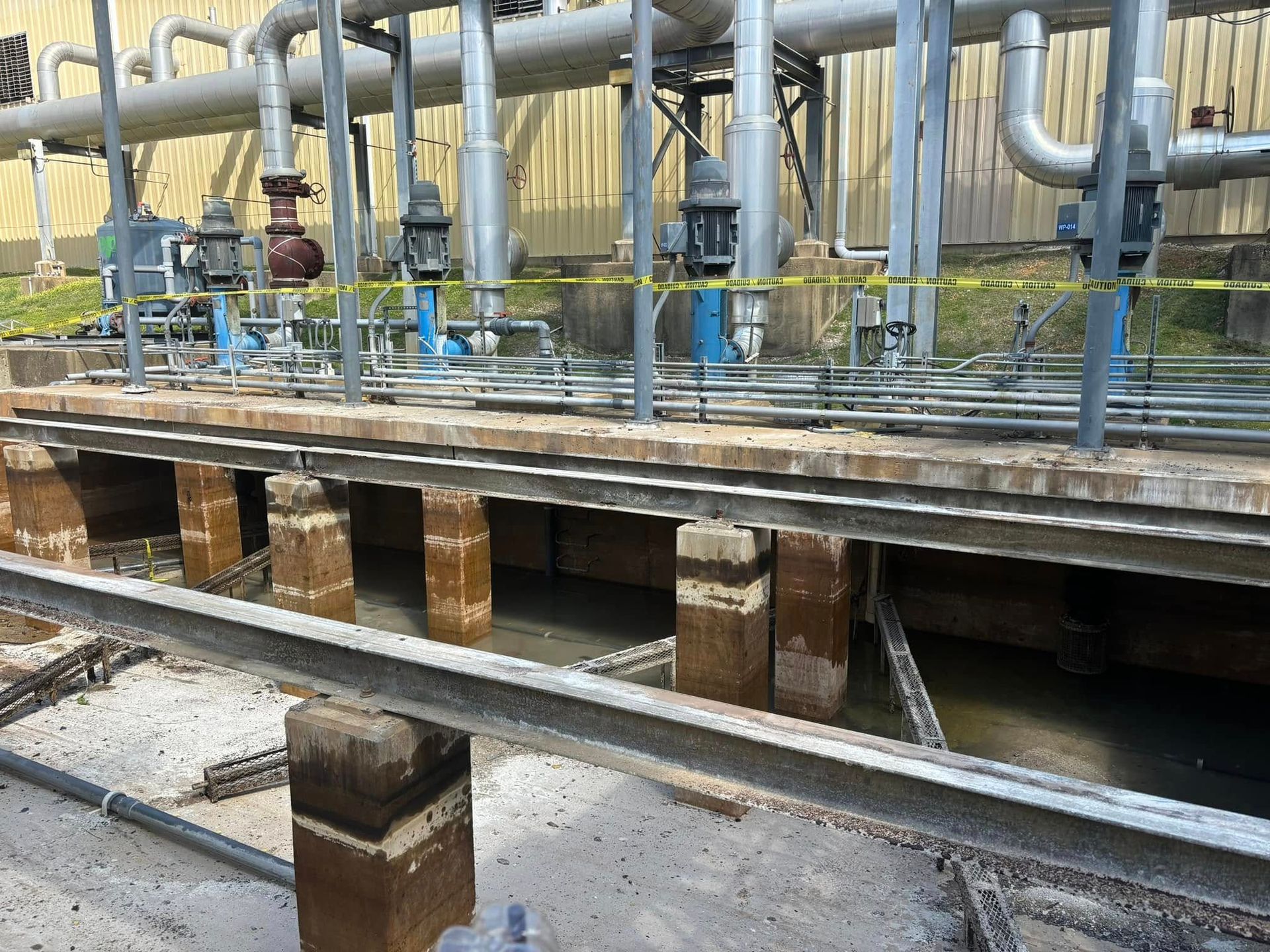 Industrial machinery with overhead pipes and blue pump stands mounted on concrete supports above a shallow water basin.