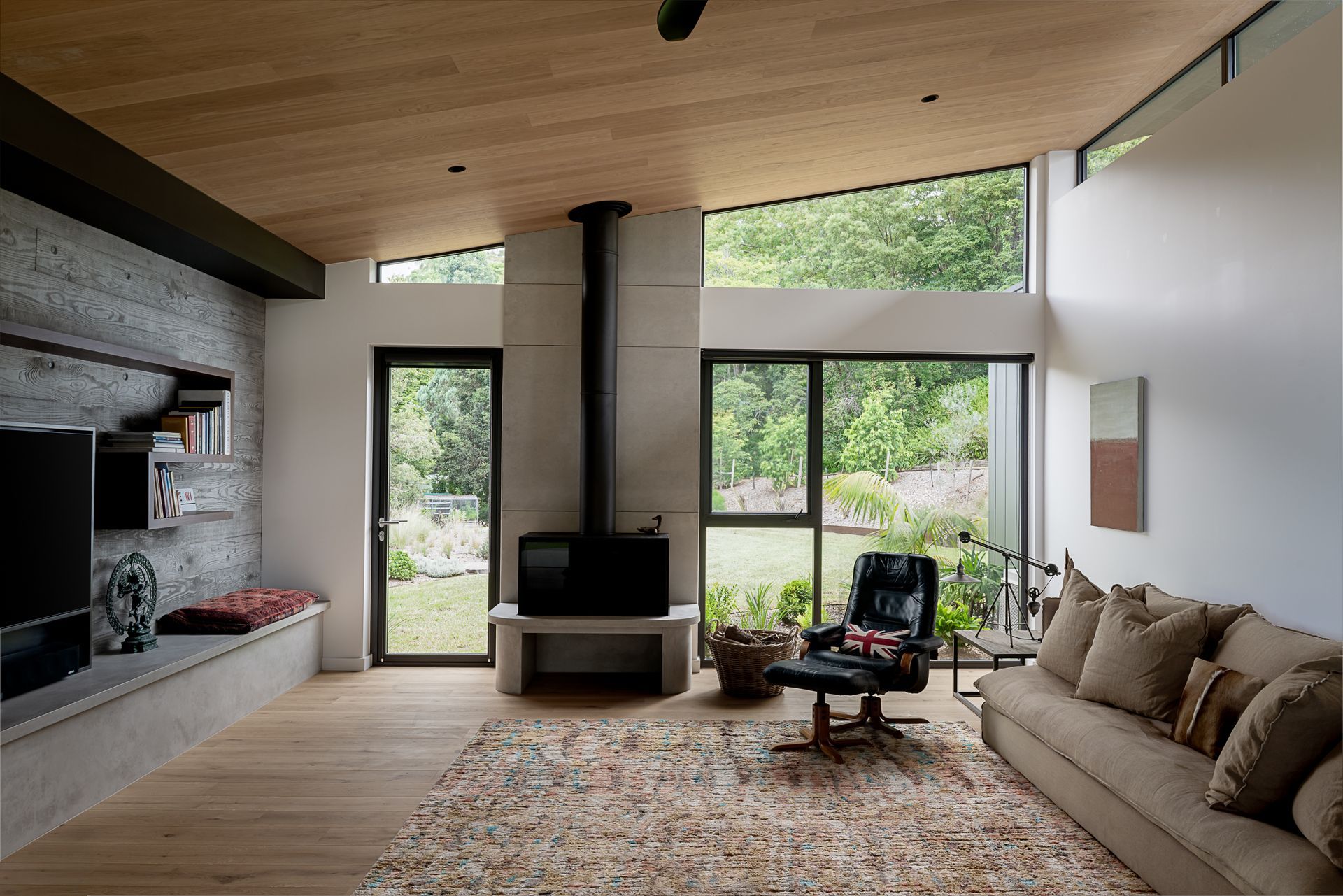 Modern living room with large windows, wood ceiling, concrete wall, and a view of the outside greenery. — Goode By Design in Unanderra, NSW