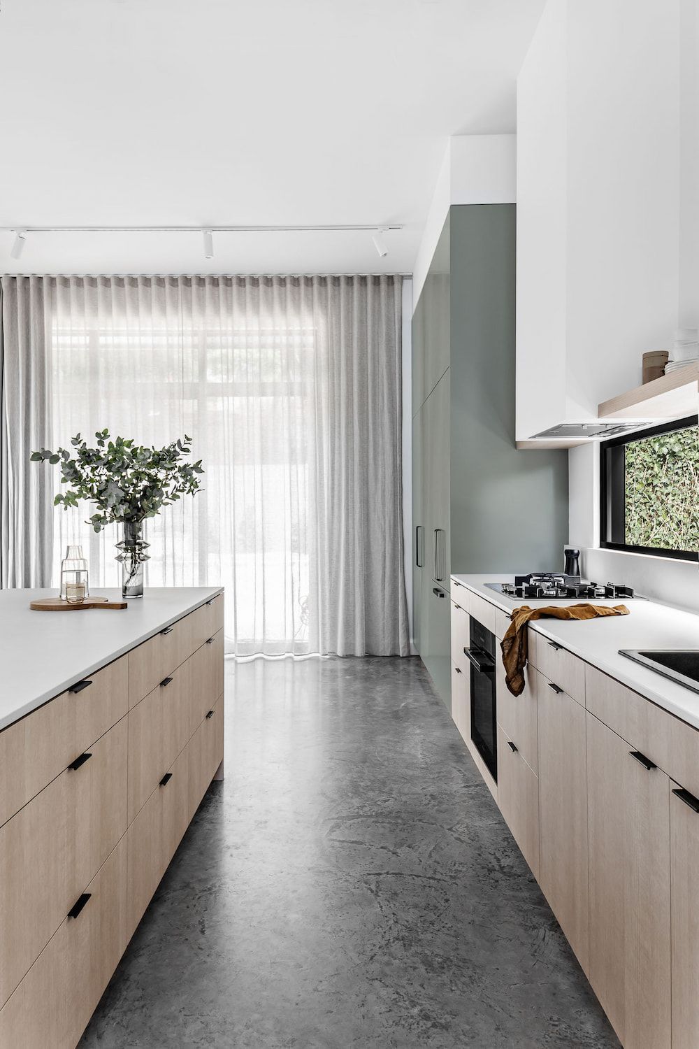 Modern kitchen with light wood cabinets, gray concrete floor, and green wall; window with lush greenery outside. — Goode By Design in Unanderra, NSW