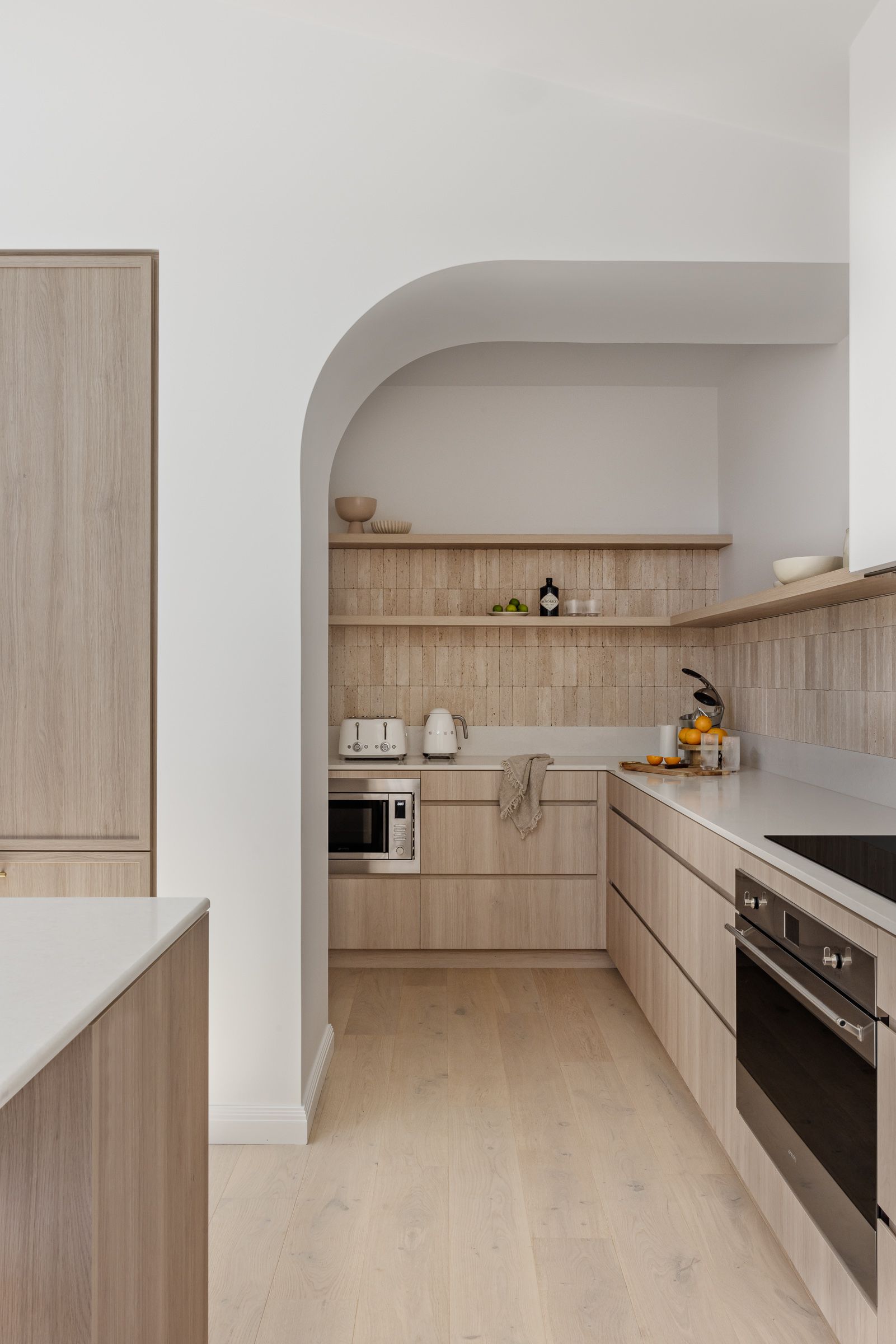 A small kitchen with light wood cabinets and floors, an arched doorway, and a built-in oven. — Goode By Design in Unanderra, NSW