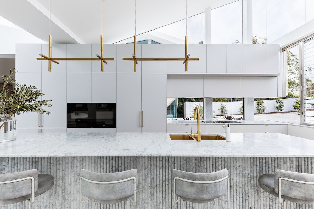 Modern white kitchen with marble island, gold fixtures, and gray bar stools. — Goode By Design in Sydney Joinery, NSW