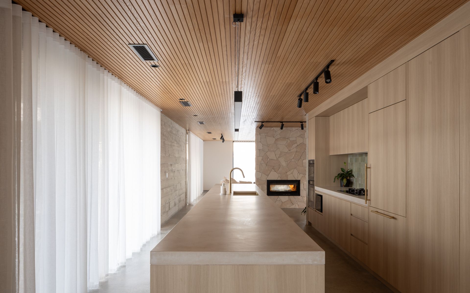 Kitchen with wooden ceiling, long island, light wood cabinets, fireplace, and sheer curtains.