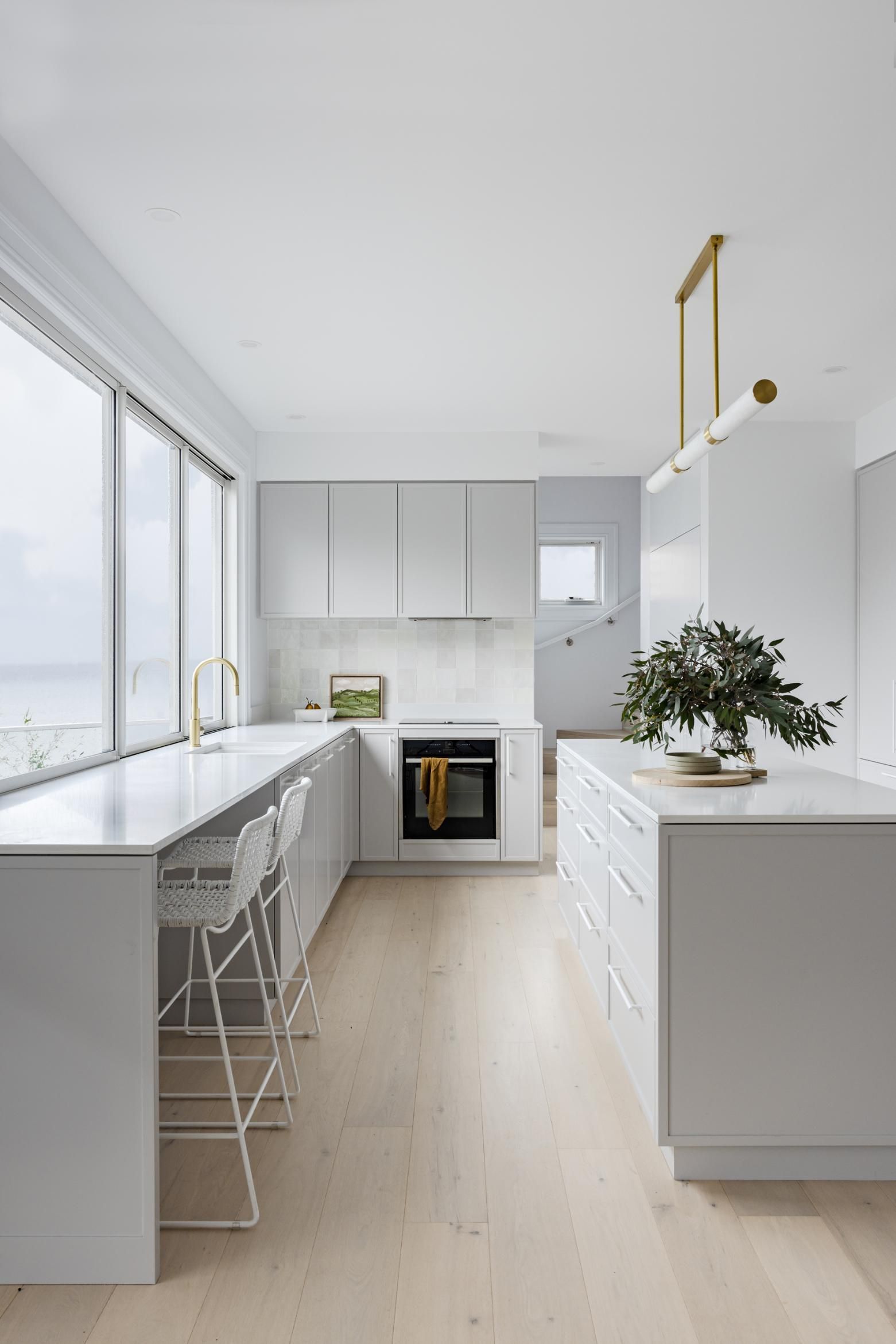 Modern white kitchen with island, seating area by window, and light wood flooring. — Goode By Design in Unanderra, NSW