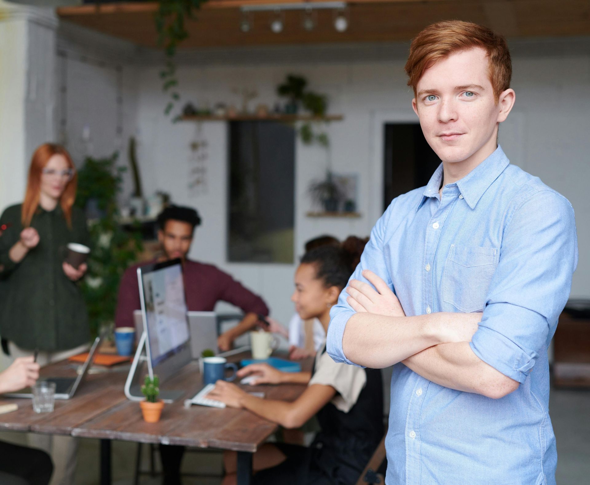 A man with his arms crossed is standing in front of a group of people in an office.