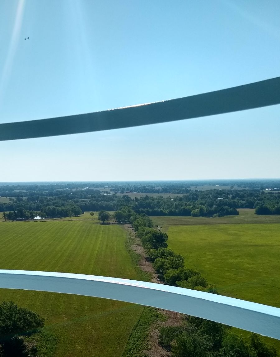 View from above of fields, trees, and a blue sky. Curved metal bands frame the scene.