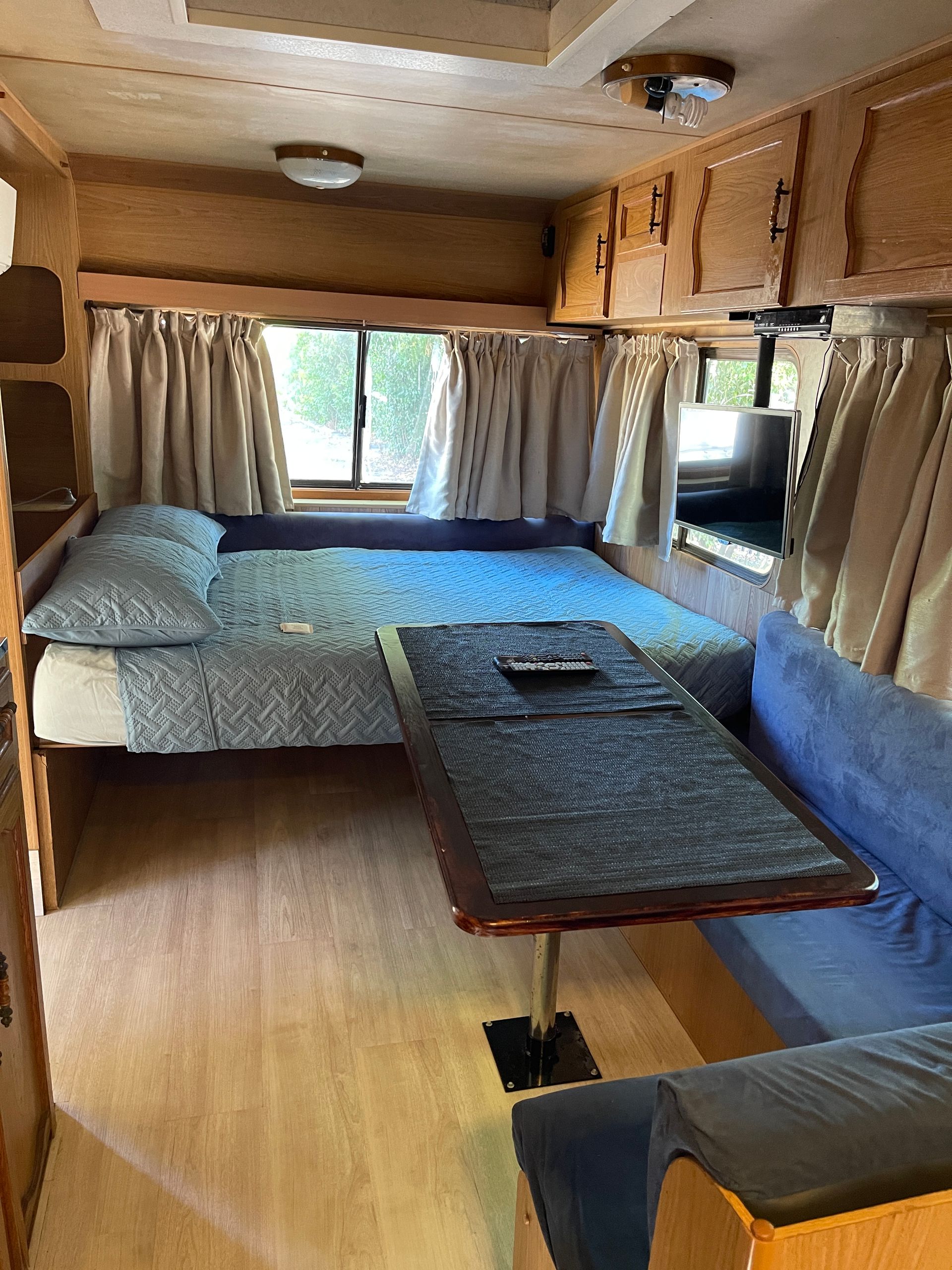 Interior of a Vintage Rv: Bed, Table, Blue Seating, Wood Cabinets, Small Window, Curtains, and a Tv — Karumba Point Holiday Tourist Park in Karumba, QLD