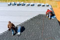 Two workers install dark gray asphalt shingles on a roof, working from bundles placed along the top edge.