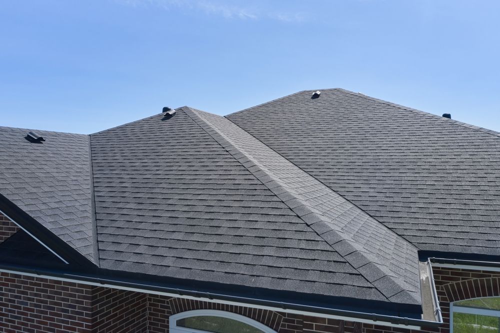 A high-angle view of a grey shingled roof with multiple peaks and vents against a bright blue sky.