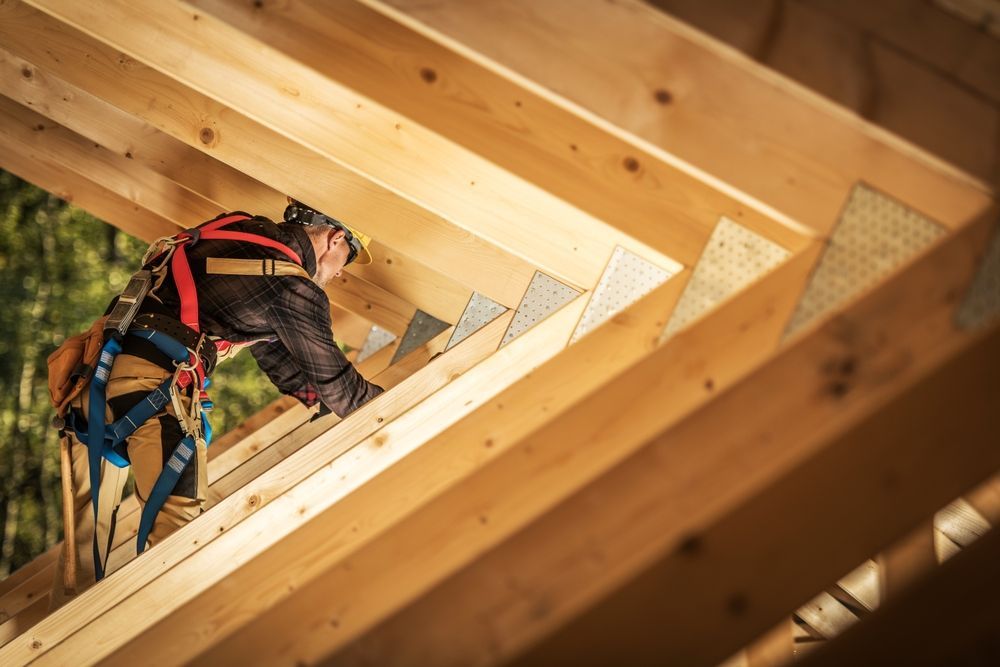 A worker wearing a safety harness and hard hat installs wooden beams on a roof frame.