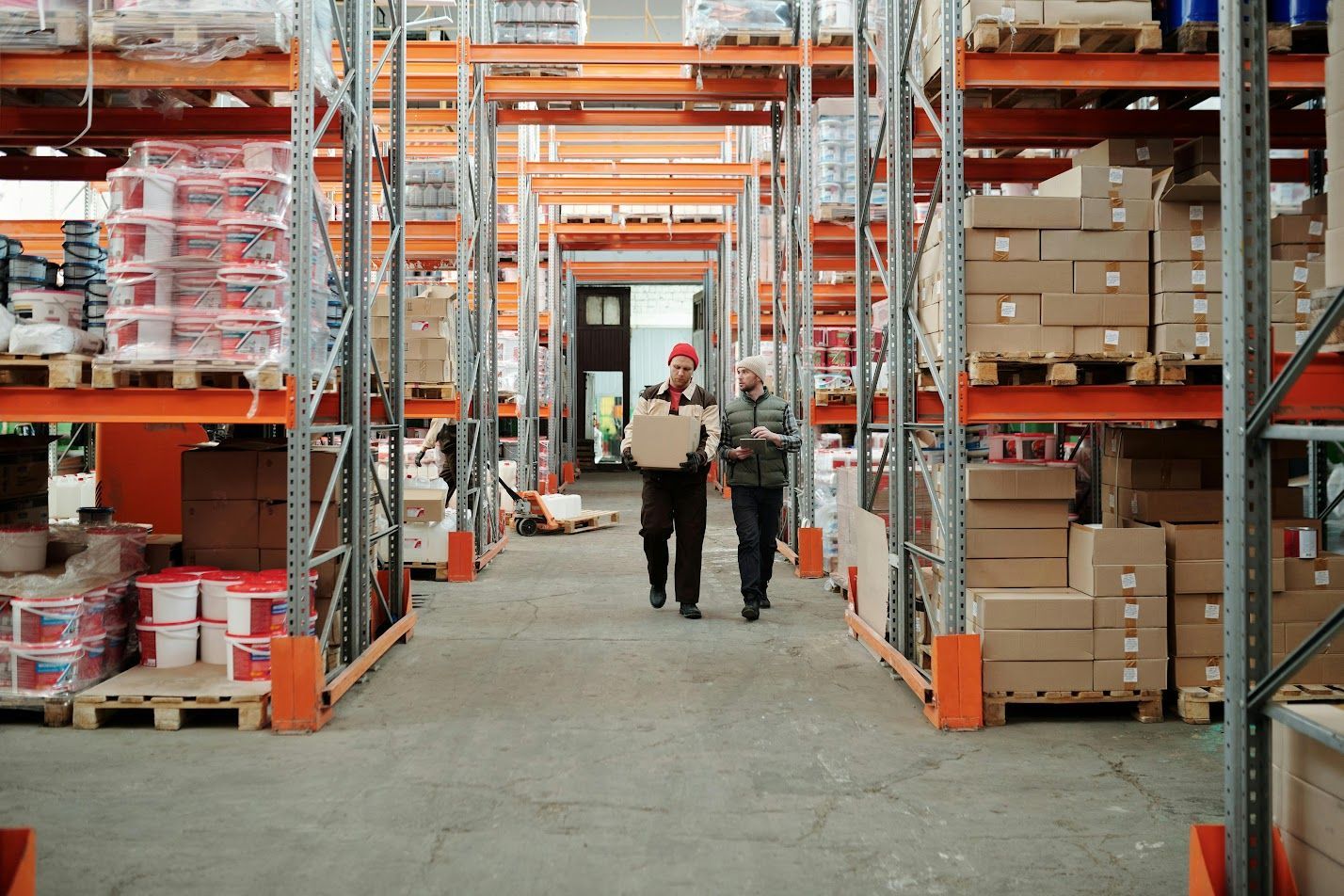 Two men are walking through a warehouse carrying boxes.